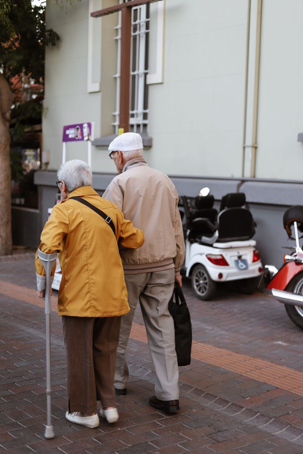 An older couple walking together outdoors, one using a cane