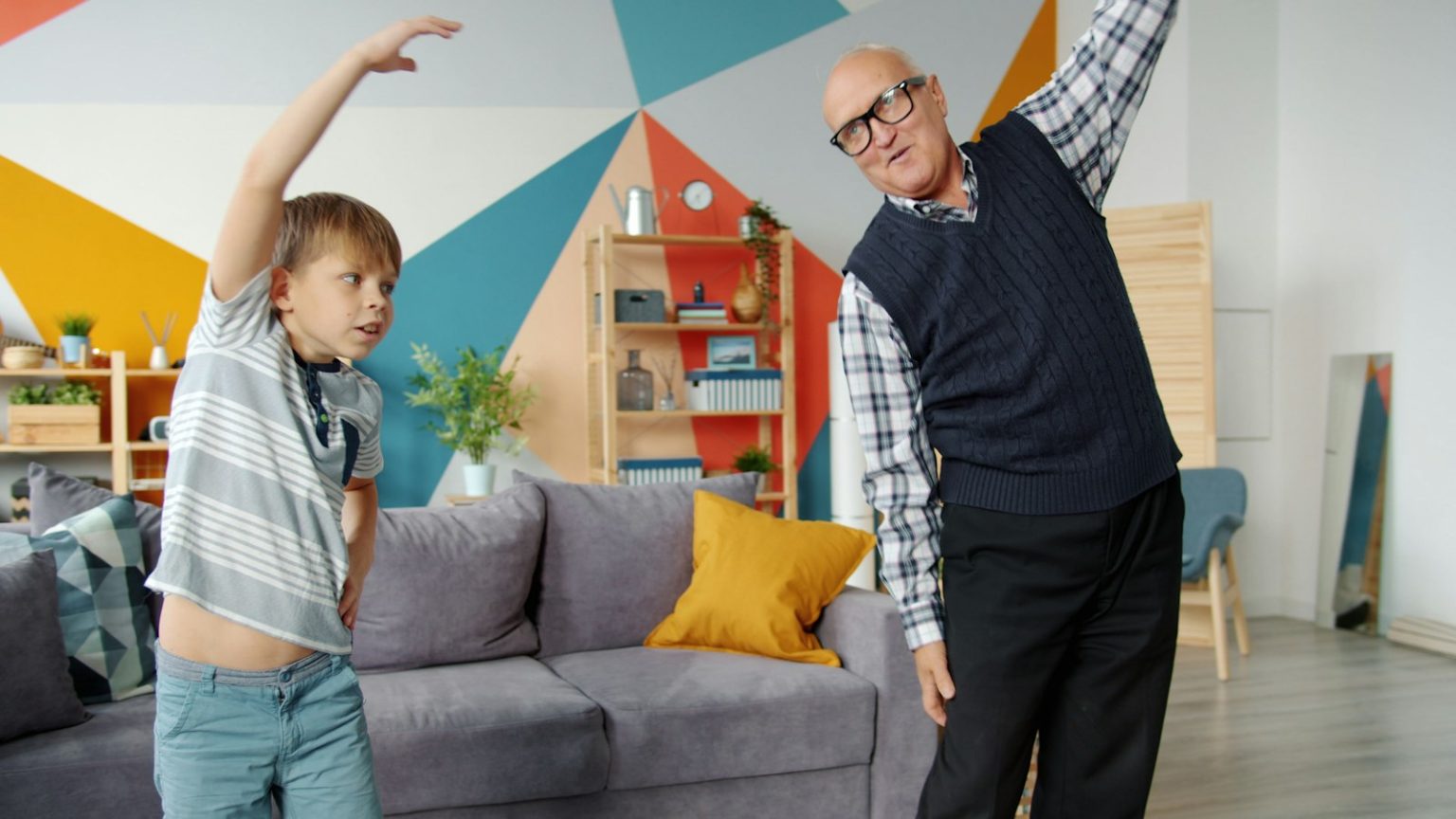 An older man doing a standing stretch at home with his grandson