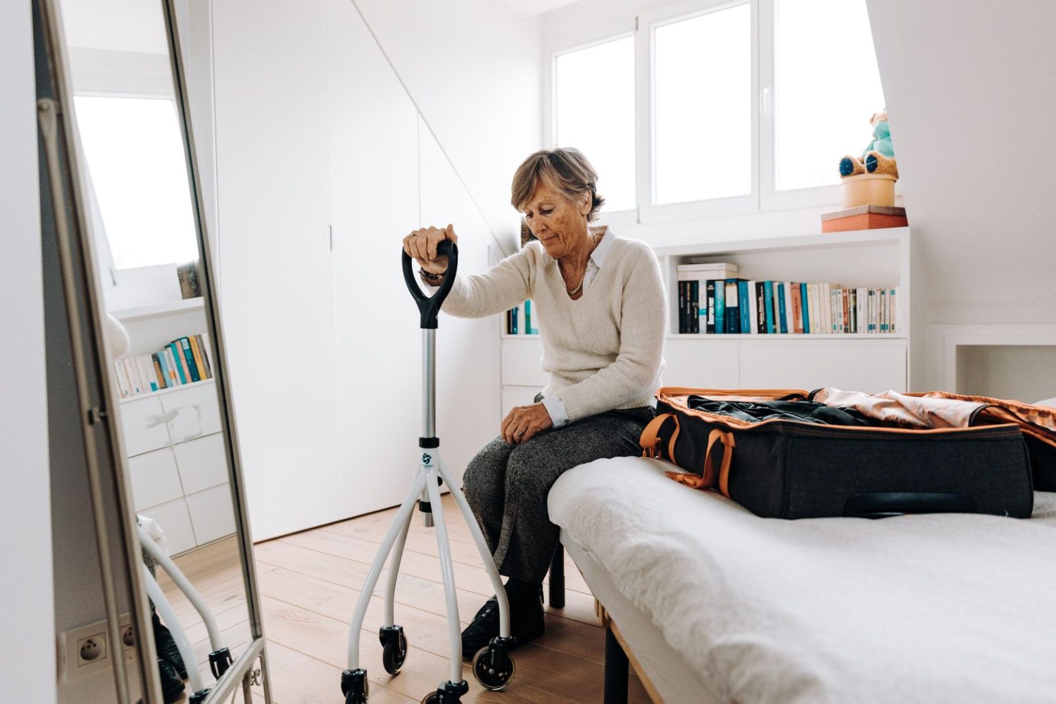 An older woman sitting on the edge of her bed with her walker beside her