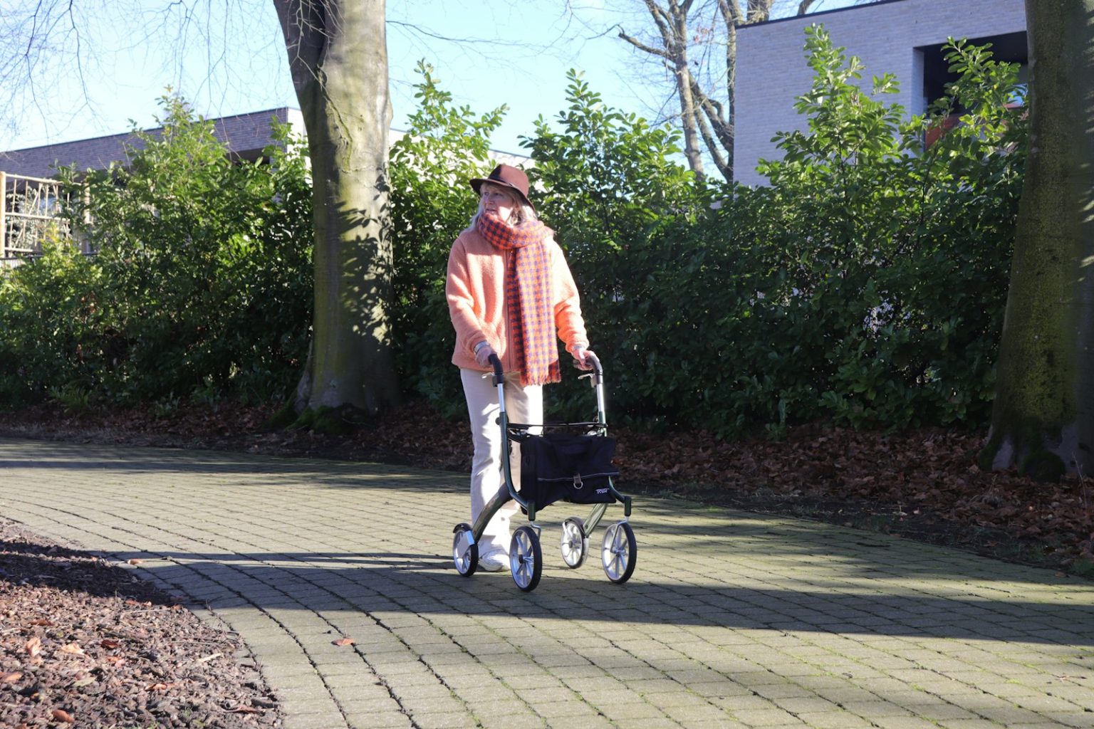 An older adult walking outdoors on a paved path with a rollator